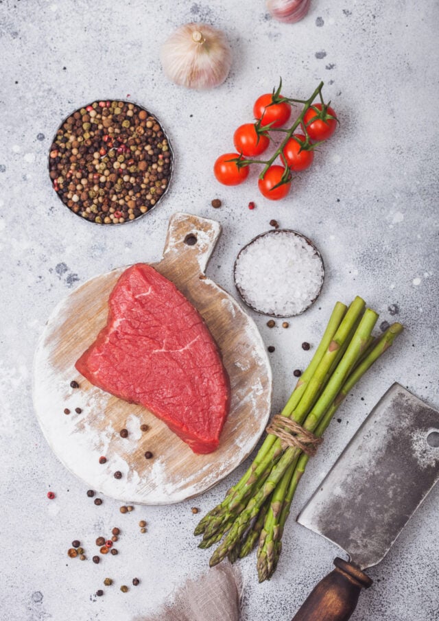 Slice of Raw Beef sirlion steak on round chopping board with tomatoes,garlic and asparagus tips and meat hatchet on light kitchen table background.