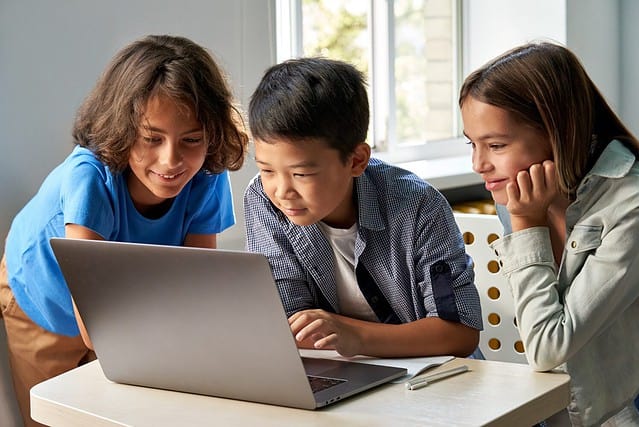 Diverse happy school kids using laptop computer together in classroom. Multicultural children junior students classmates learning online elementary education program class gathered at desk.