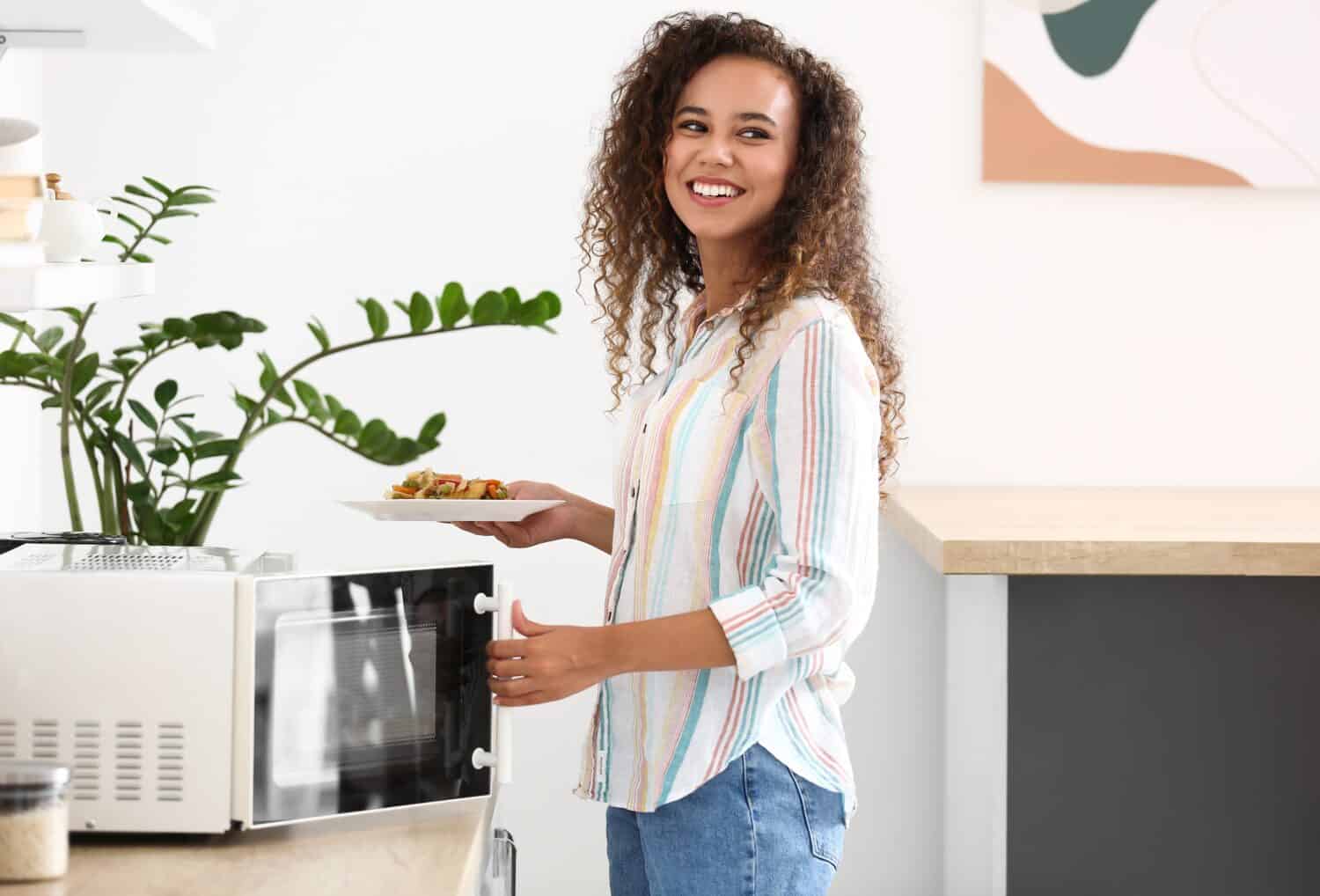 Young African-American woman putting plate with food into microwave oven in kitchen