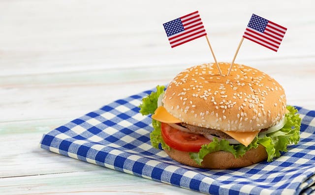 Closeup studio shot delicious tasty minced beef meat cheeseburgers fast food with two small United States of America national flag pinned on bread bun placed on plaid napkin on old retro wooden table.