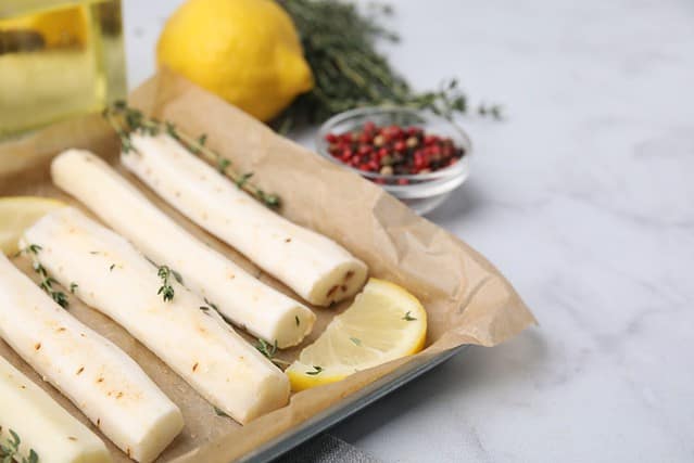 Baking tray with raw salsify roots, lemon and thyme on white marble table, closeup. Space for text