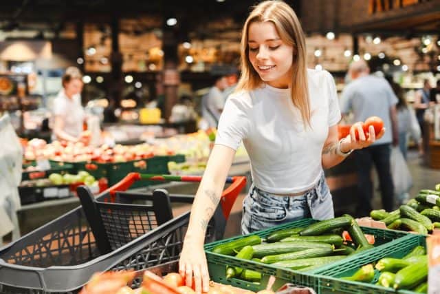 Young woman happily shops for fresh produce in a grocery store, selecting tomatoes and cucumbers for a healthy lifestyle. The vibrant vegetables stand out against a blurred background of shoppers