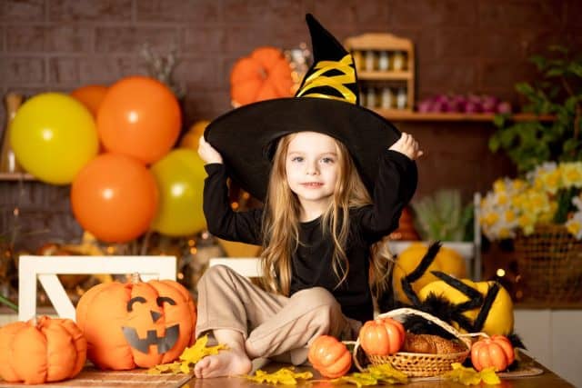 halloween, a cute smiling child girl in a witch costume in a dark kitchen with pumpkins and a big spider and festive decor during the Halloween celebration rejoices