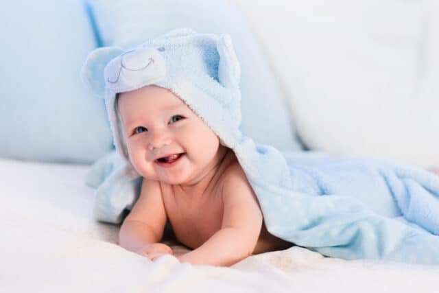 Baby boy wearing diaper and blue towel in white sunny bedroom. Newborn child relaxing in bed after bath or shower. Nursery for children. Textile and bedding for kids. New born kid with toy bear.