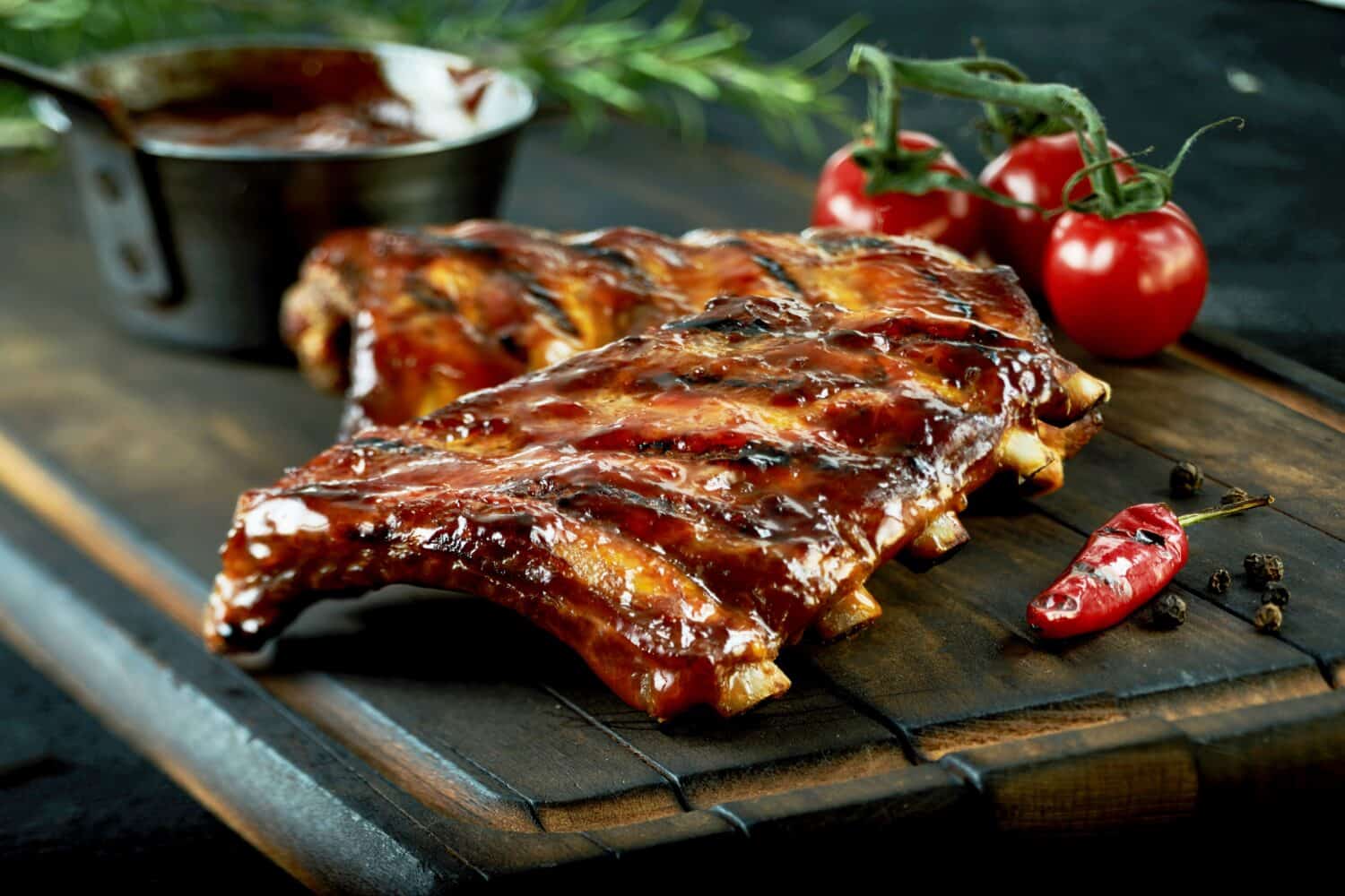Spicy hot grilled spare ribs from a summer BBQ served with a hot chili pepper and fresh tomatoes on an old vintage wooden cutting board