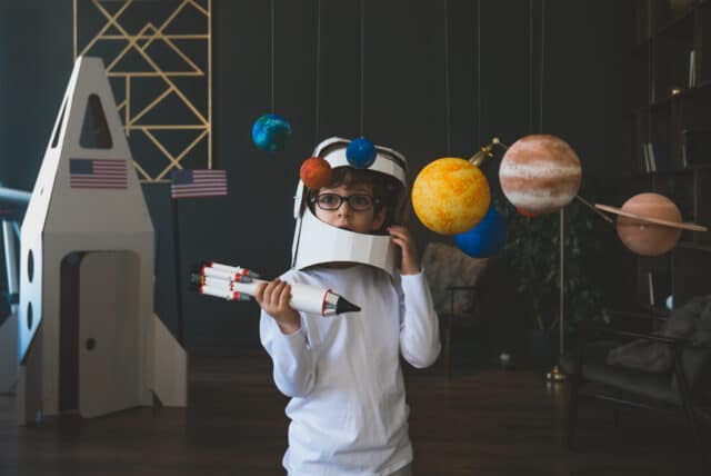 Cute little boy wearing cardboard astronaut helmet flying toy rocket through planets, cardboard spaceship rocket in the background